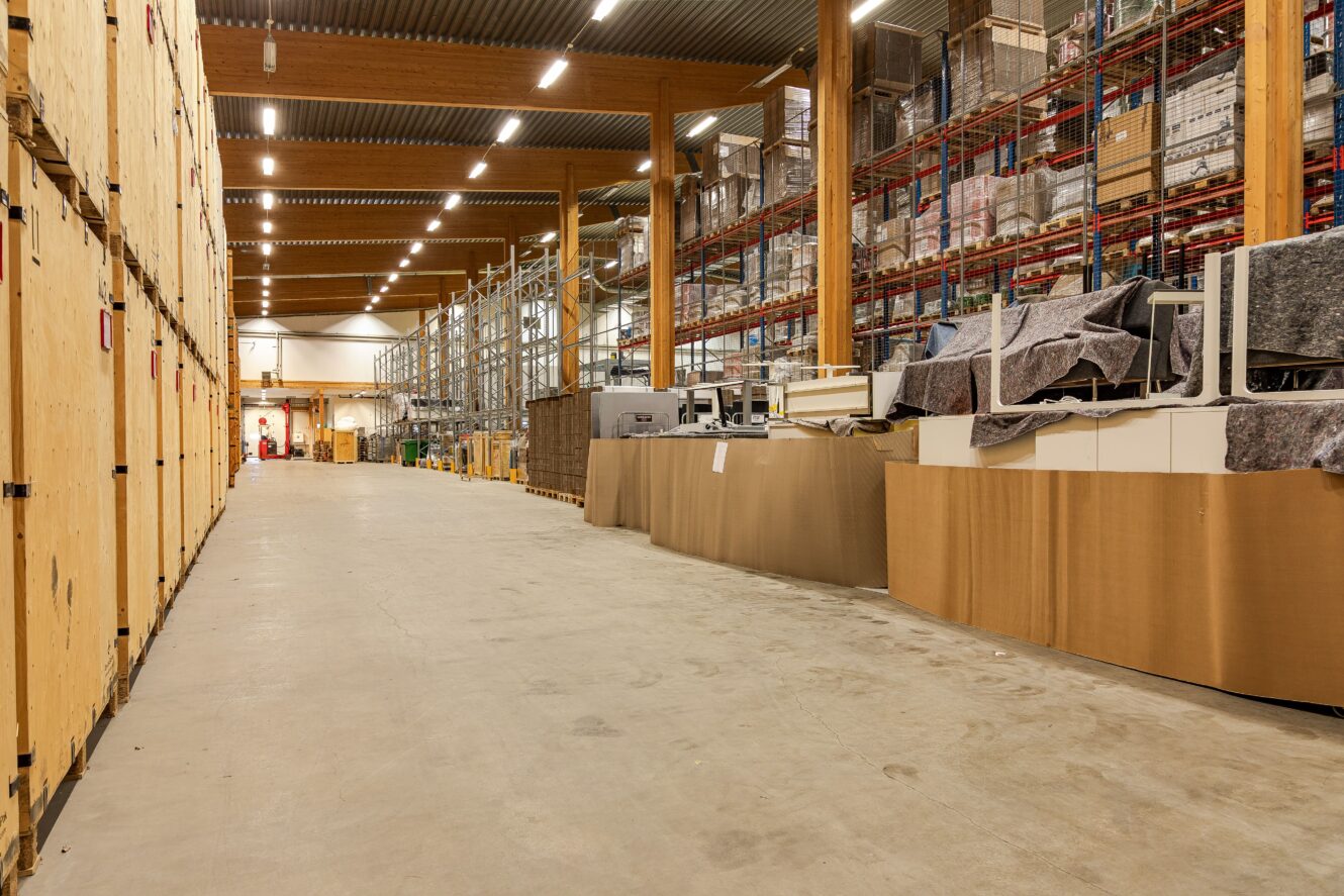 Stor lagerlokal med hyllor fyllda med lådor och varor, trälådor till vänster och diverse förpackade föremål till höger. Utrymmet är väl upplyst och organiserat.