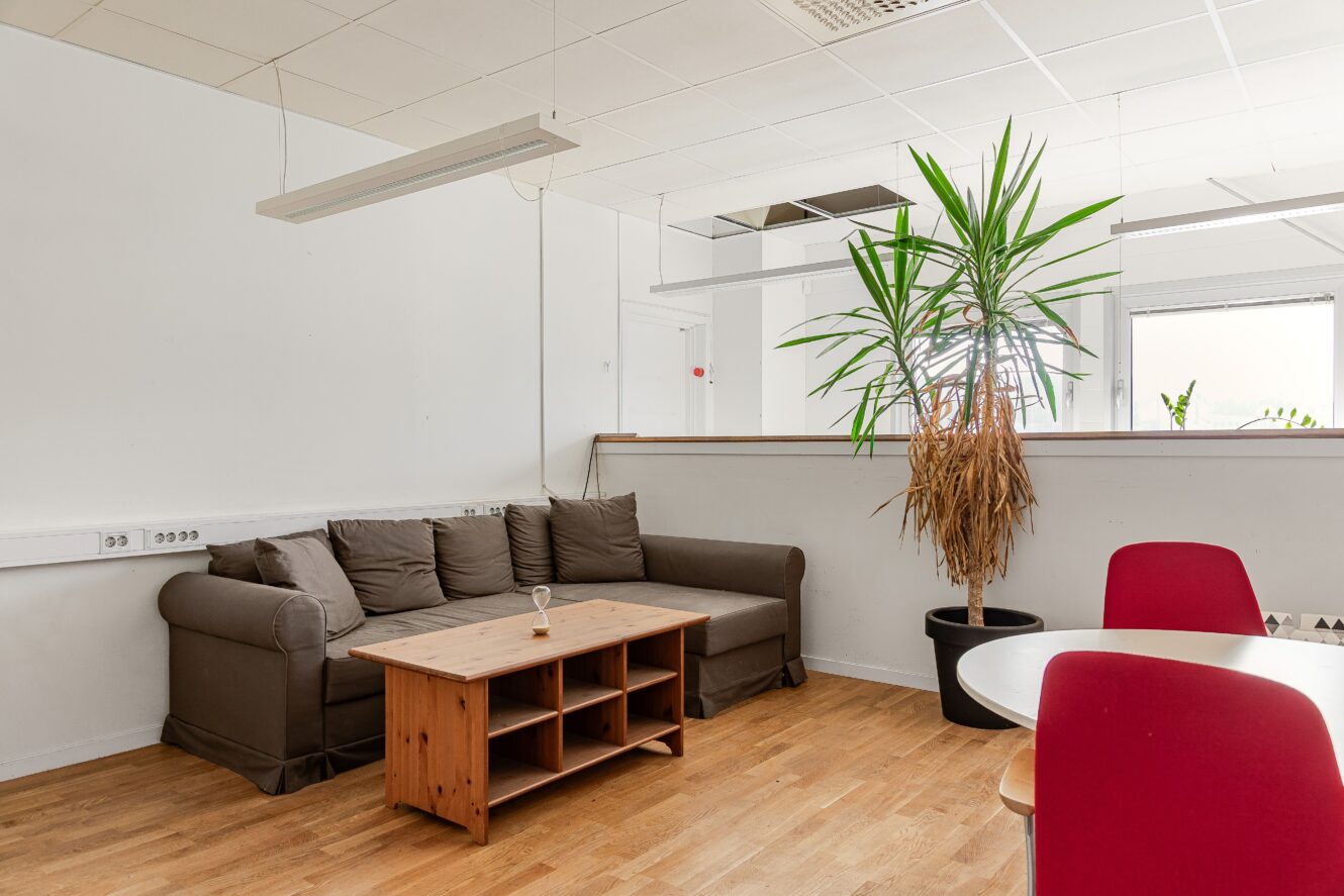 Ett litet kontorsloungeområde med en brun soffa, soffbord i trä, krukväxt och röda stolar runt ett bord på ett trägolv.