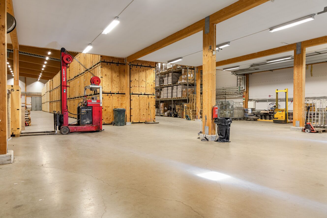 Rymlig lagerlokal med stödbalkar i trä, stora trälådor, gaffeltruckar, pallar och industrihyllor.