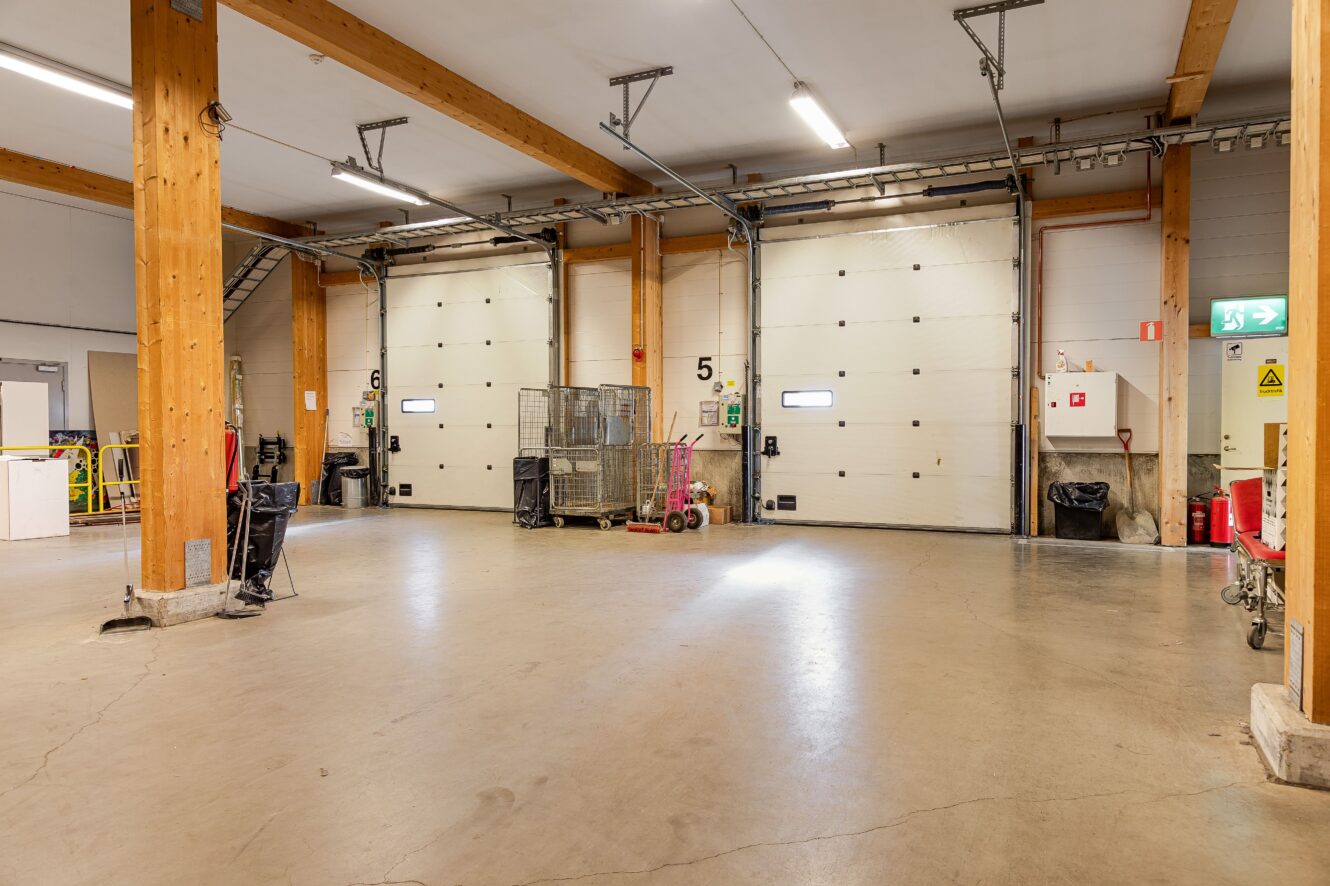 Interiör från en lastplats i ett lager med betonggolv, träpelare, numrerade takskjutportar, vagnar och synlig säkerhetsutrustning.