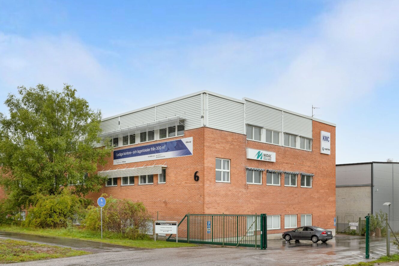 En tvåvånings kontorsbyggnad i tegel med flera företagsskyltar, en gated entrance, parkerade bilar och ett träd på vänster sida under en blå himmel.