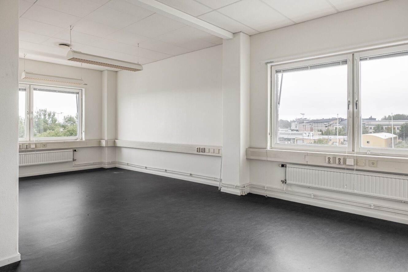 Tomt rum med vita väggar, stora fönster, svart golv och taklampor; utsikten visar träd och byggnader under en molnig himmel.