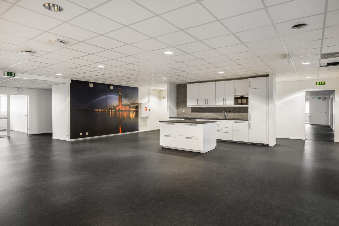 Ett modernt, tomt kontorsutrymme med vita skåp och en ö i mitten, mörkt golv och en stor väggmålning av en fyr i solnedgången.