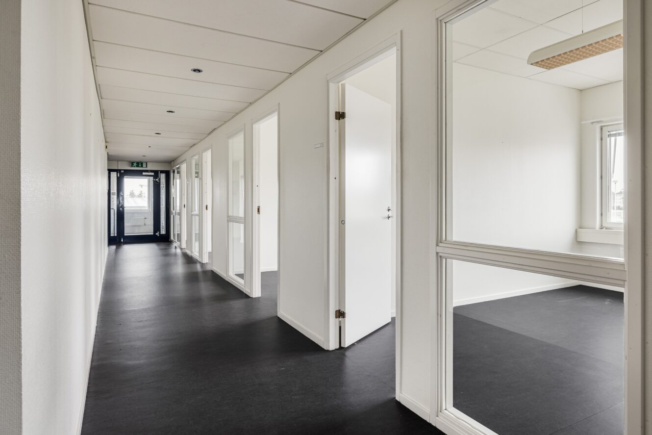 En ren, tom korridor med vita väggar och dörrar som leder till omöblerade rum med stora fönster och stark takbelysning.