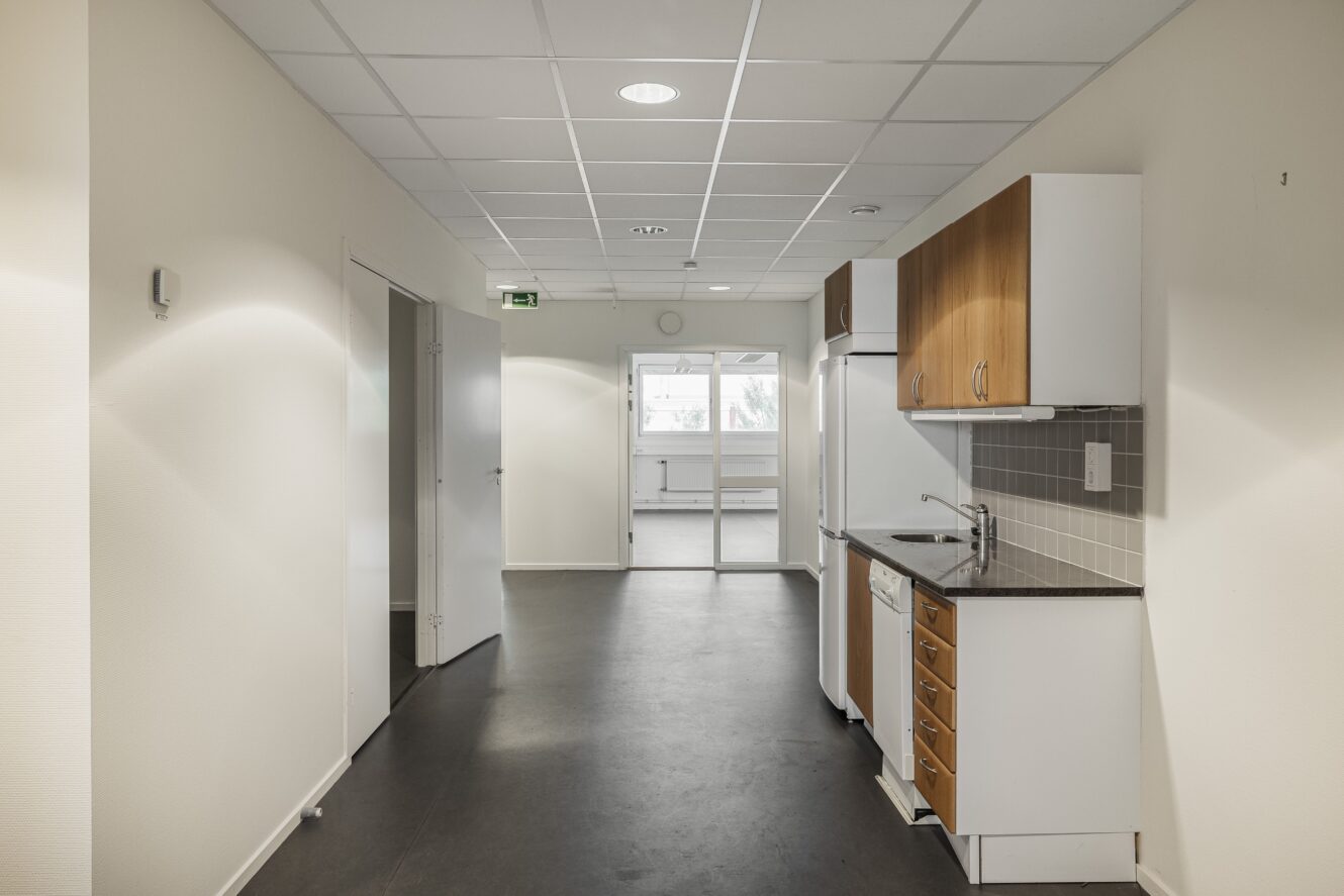 Ett litet pentry med skåp i trä och vitt, diskho och diskmaskin ligger i en hall med vita väggar och takplattor. En dörr leder till ett ljust rum i bakgrunden.