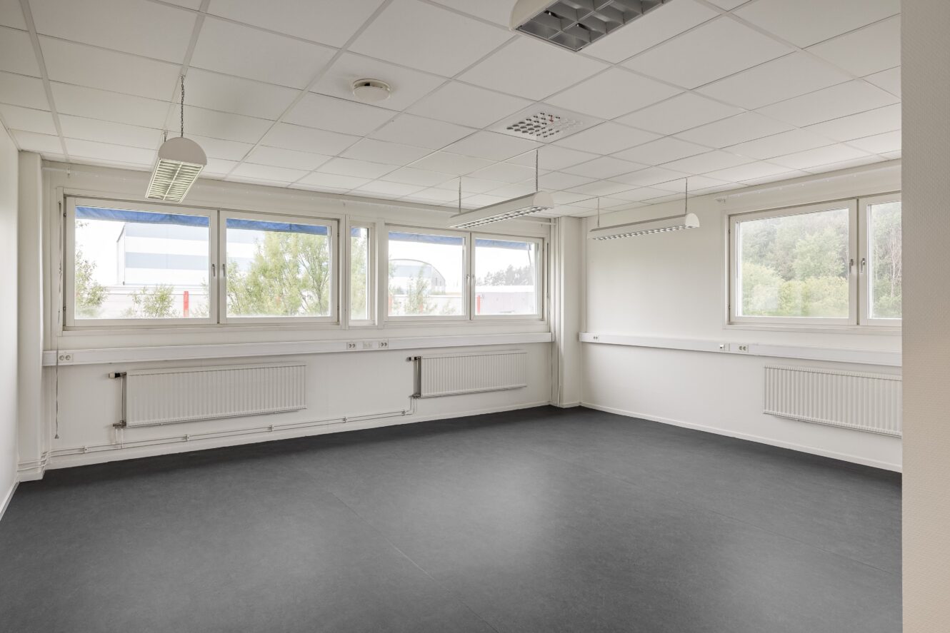 Ett tomt, omöblerat rum med vita väggar, stora fönster, ett vitt tak med lysrör och ett mörkt golv.
