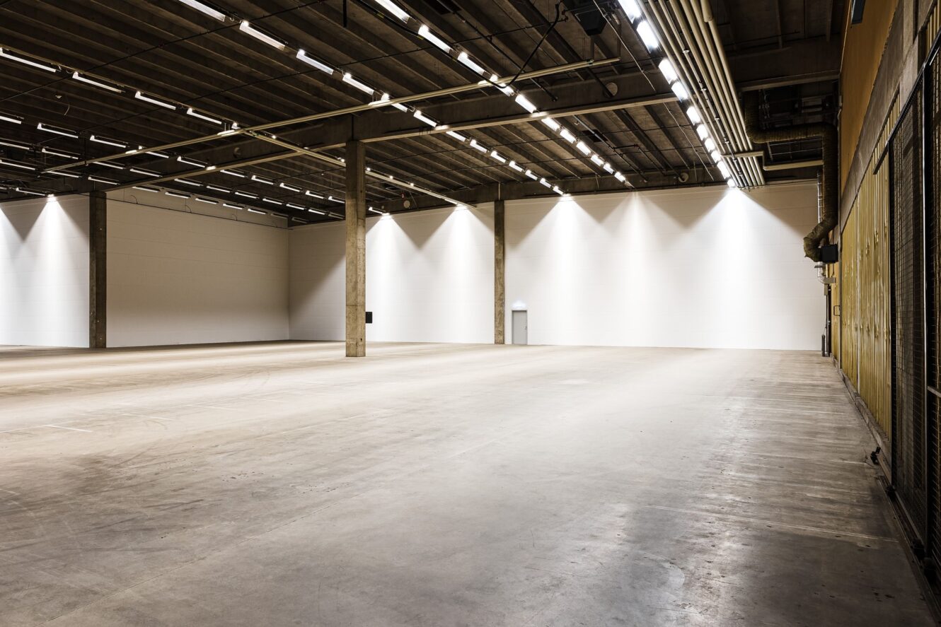 Stor, tom industrilokal med högt i tak, betonggolv, synliga bjälkar och stark takbelysning. En enda dörr syns på den bortre vita väggen.