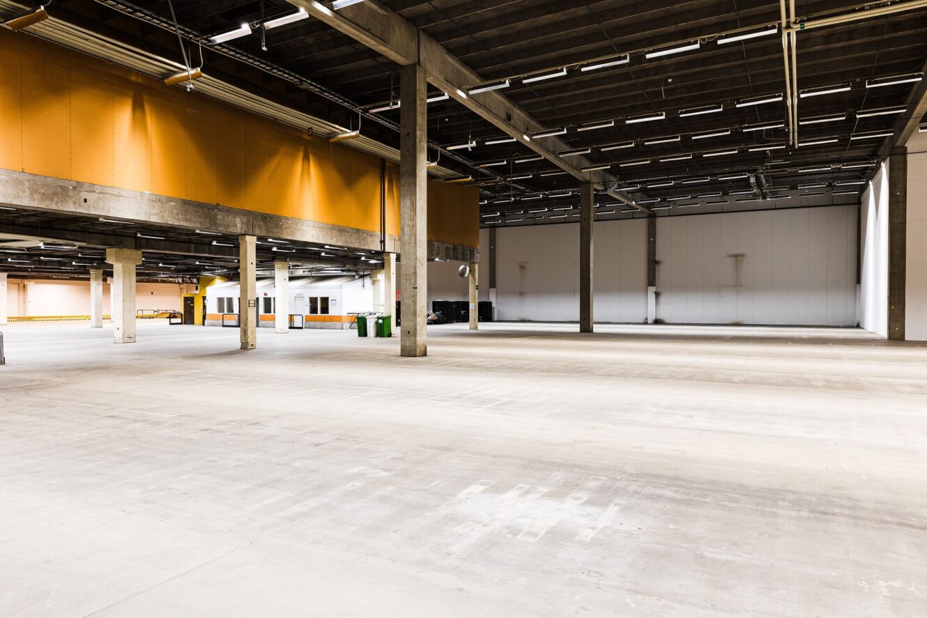 Ett stort, tomt industrilager med högt i tak, synliga bjälkar och betonggolv, upplyst av lysrör.