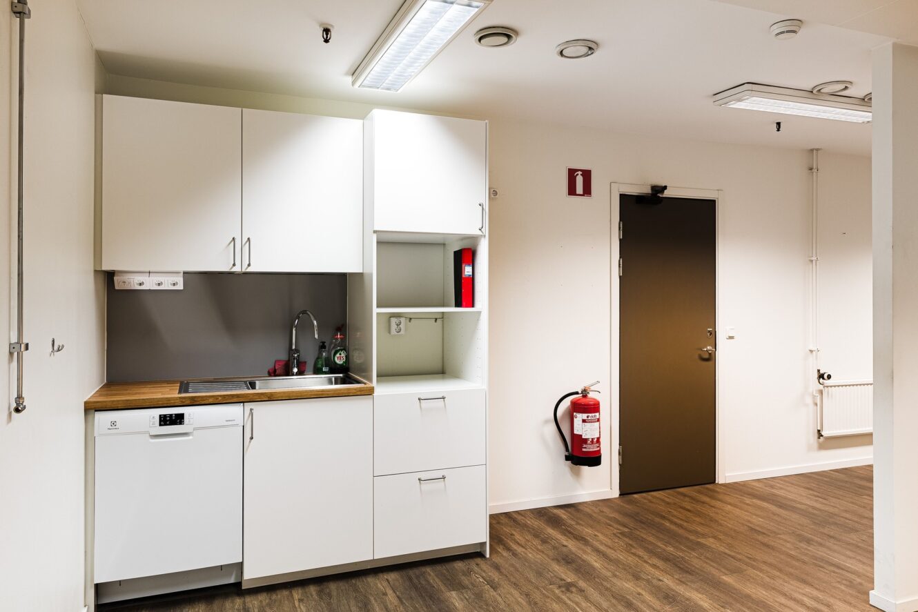 Ett litet pentry med vita skåp, diskho, diskmaskin och brandsläckare monterat nära en brun dörr på ett trägolv.