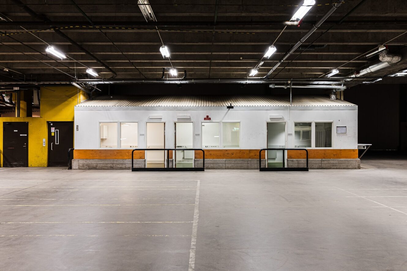 En liten, vit kontorsbyggnad med flera fönster inuti en stor, tom industrilokal med betonggolv och takbelysning.