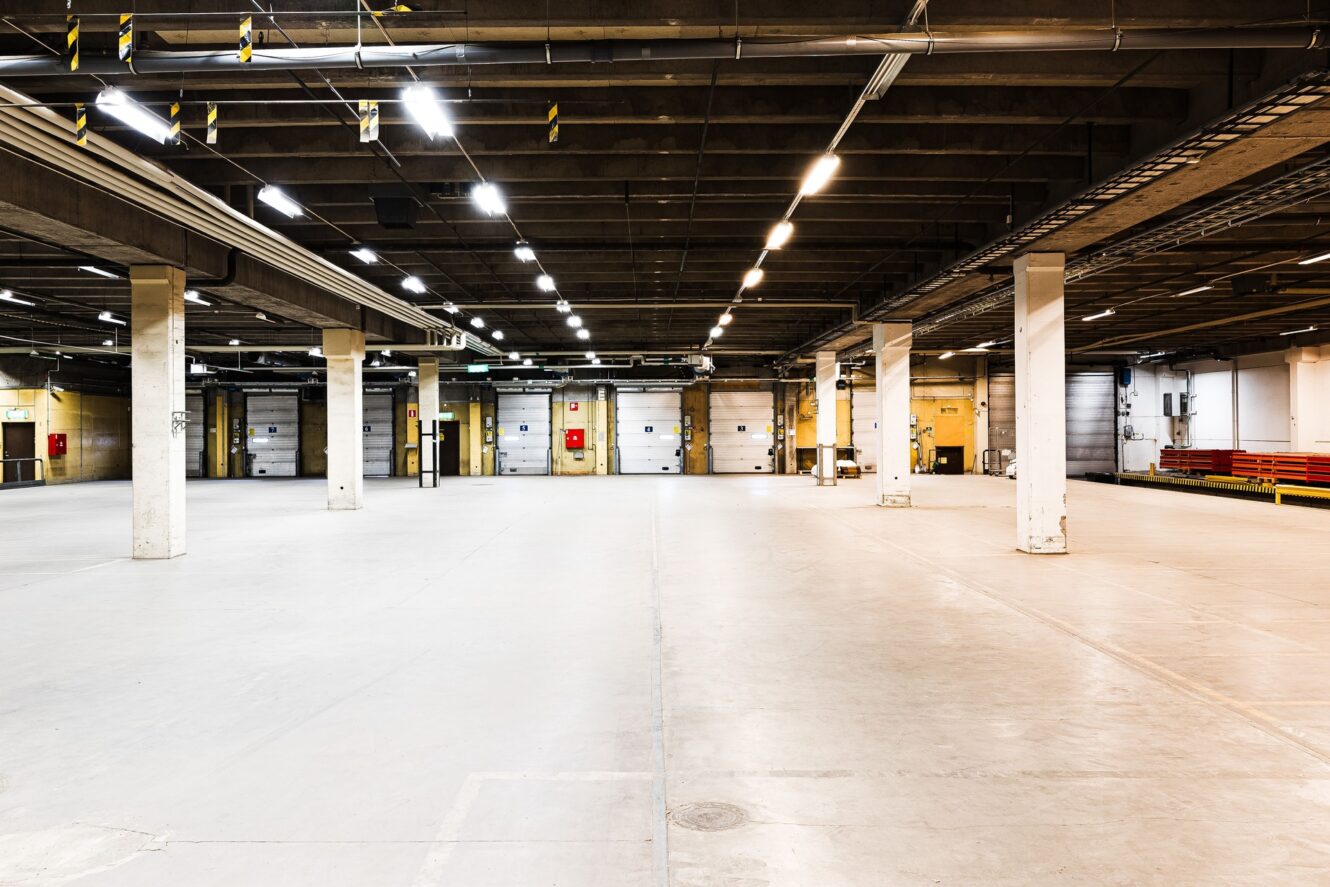 Stor, tom lagerlokal med betonggolv, högt i tak, synliga balkar och flera pelare under fluorescerande belysning. Lastkajdörrar är synliga längs den bakre väggen.