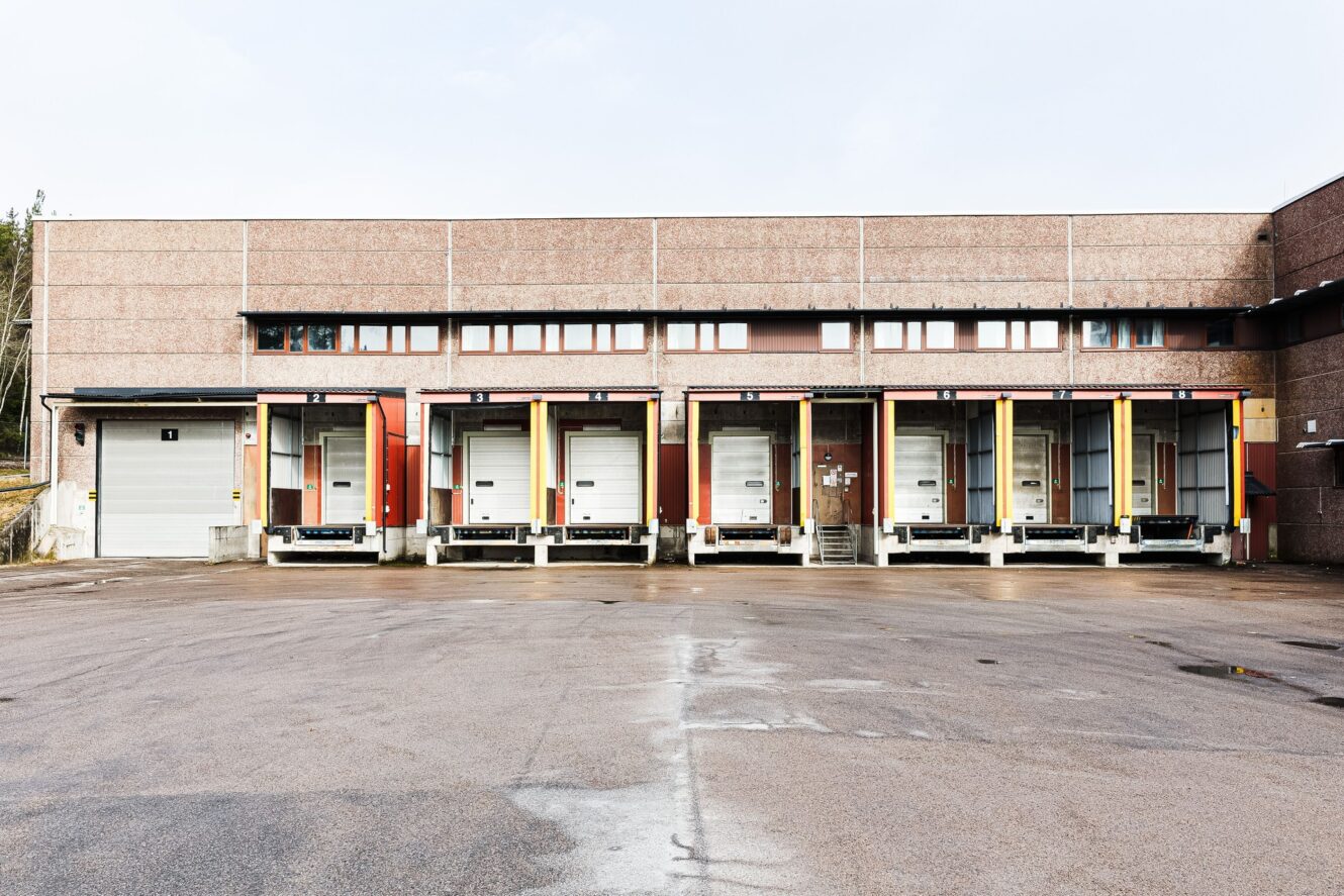 En rad med sex lastkajer med stängda dörrar vid ett lager, sett från parkeringsplatsen en molnig dag.