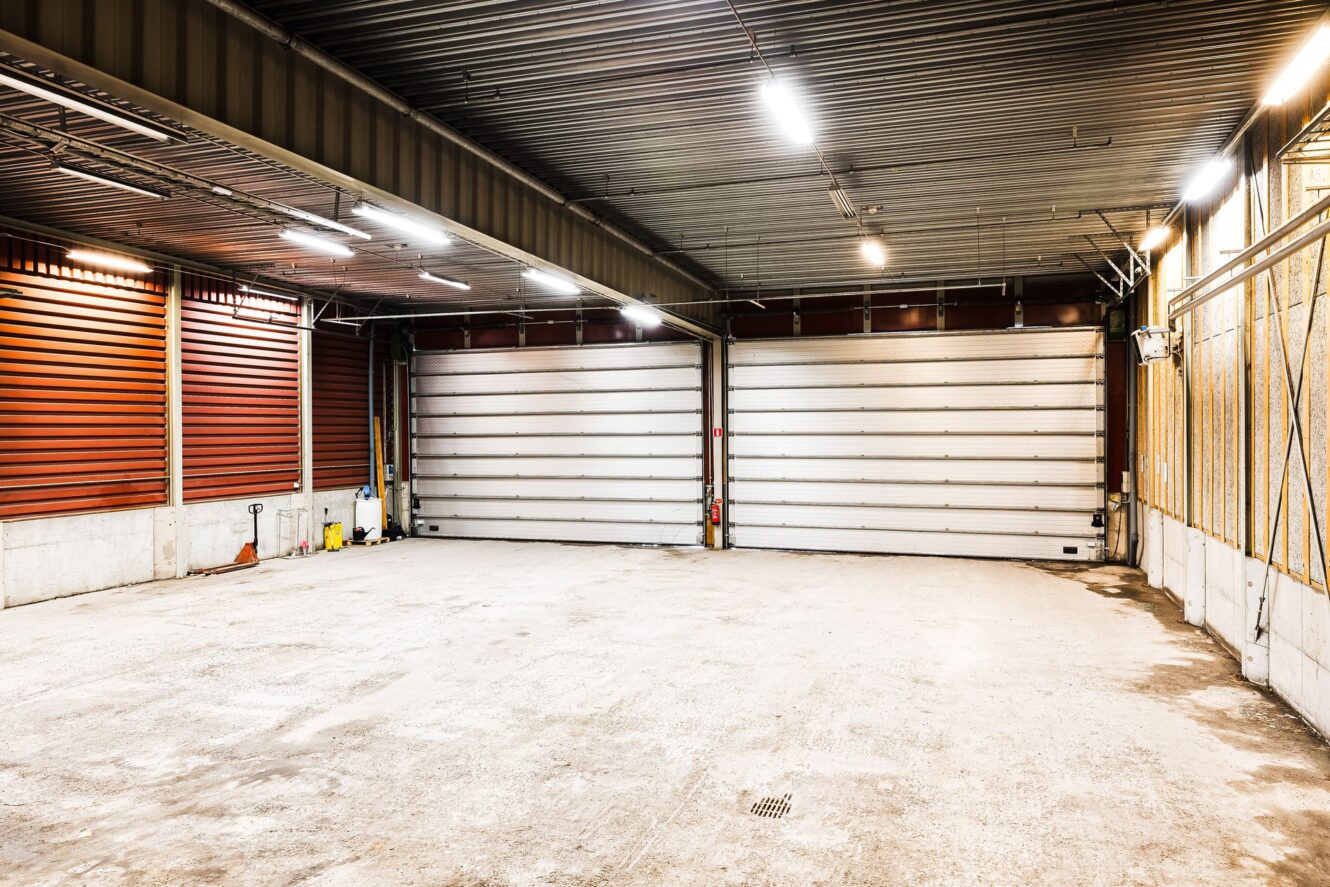 Stort, tomt industrigarage med två rullportar, betonggolv, metalltak, lysrör och städmaterial mot ena väggen.