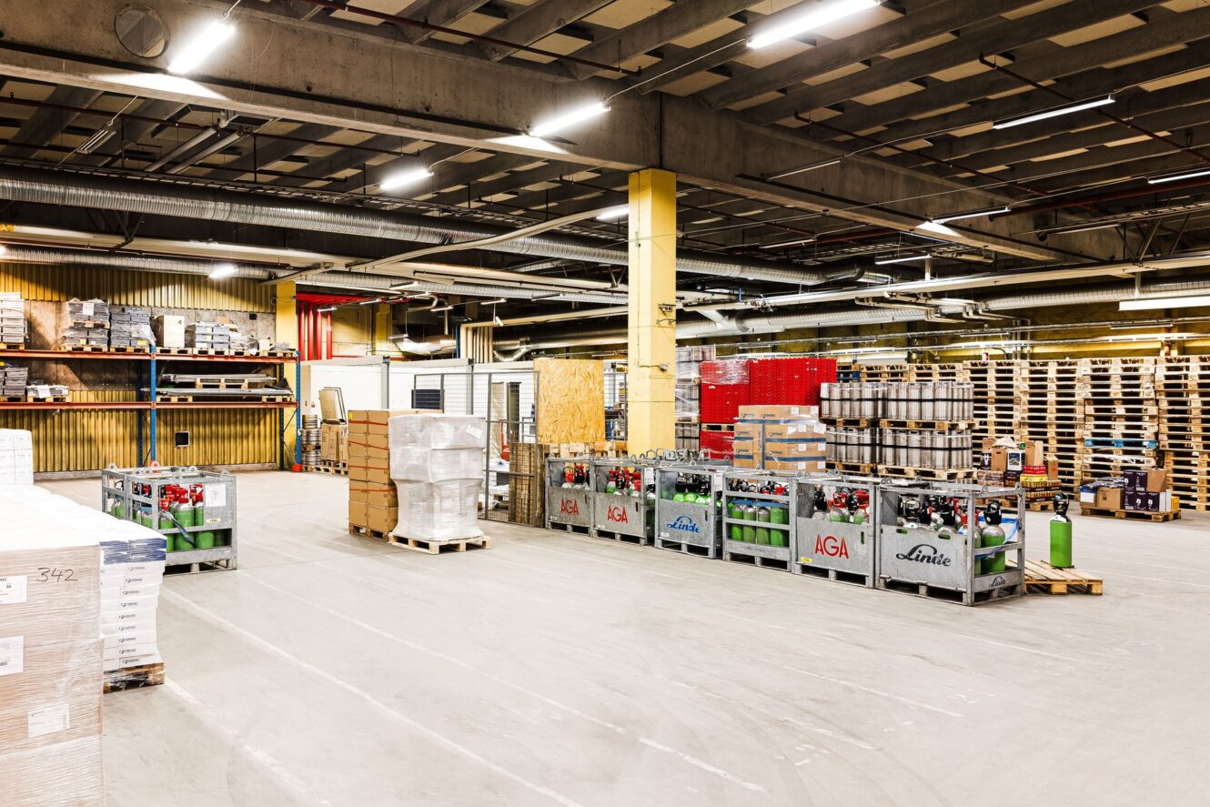 Industriell lagerlokal med staplade pallar, gasflaskor och olika varor på metallställ under lysrörsbelysning.