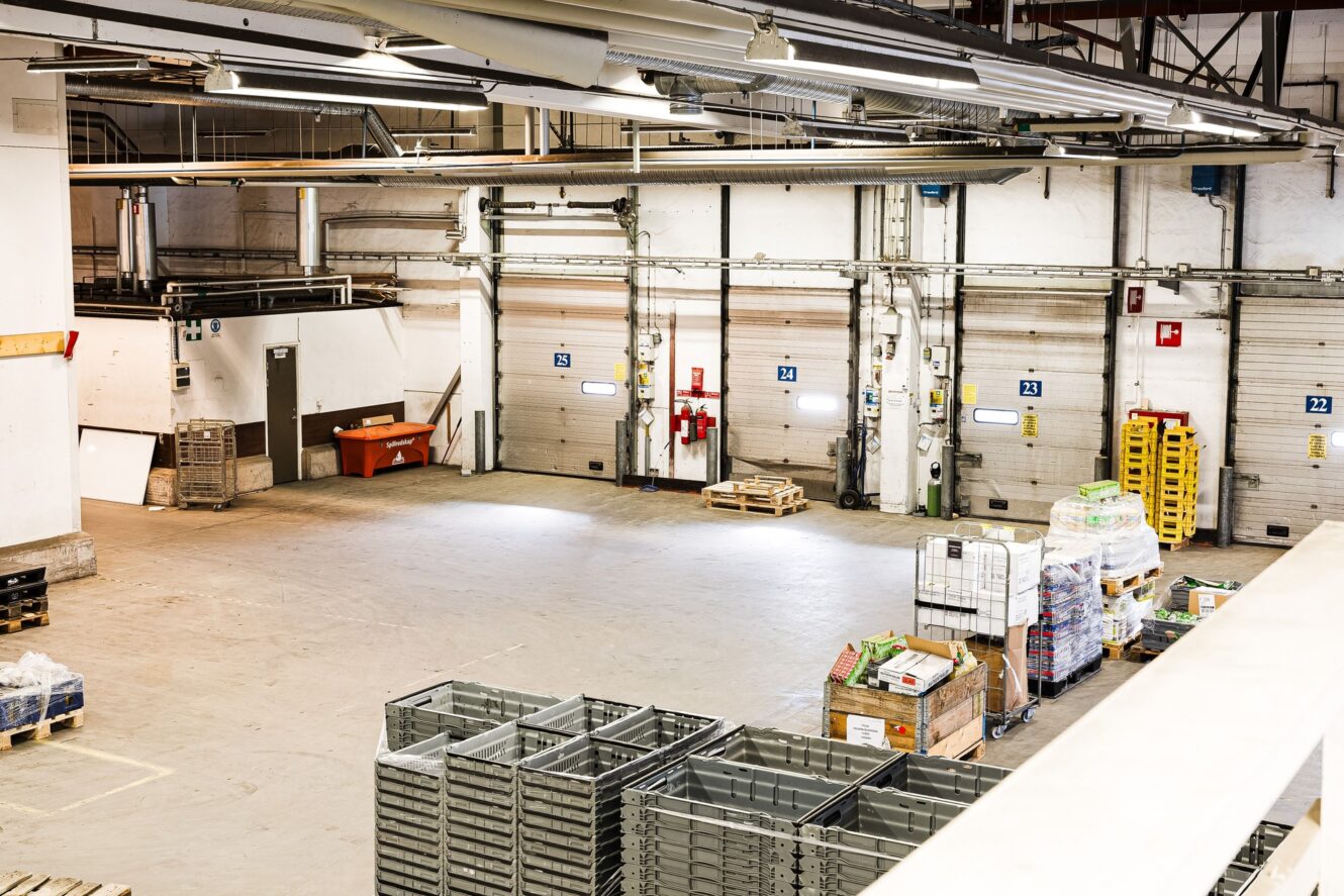 Interiör från en lagerlokal med staplade metallbackar, pallar med varor och flera lastbryggor längs bakväggen. Utrymmet är starkt upplyst och i stort sett tomt.