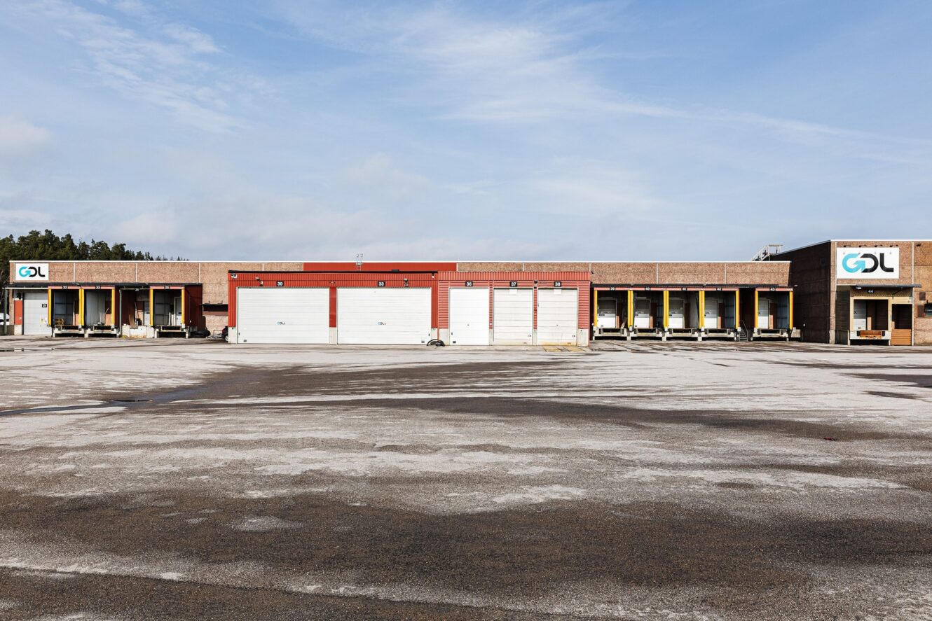 En lagerlokal med flera lastkajer och garageportar, två skyltar med texten GDL på vardera sidan och en tom parkeringsplats framför.