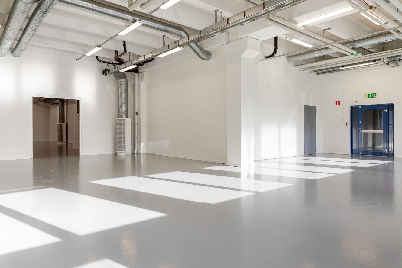 Stort, tomt industrirum med vita väggar, grått golv, synliga rör, öppen dörröppning, grå dörr och blå dubbeldörrar. Solljus strömmar in genom fönster på golvet.