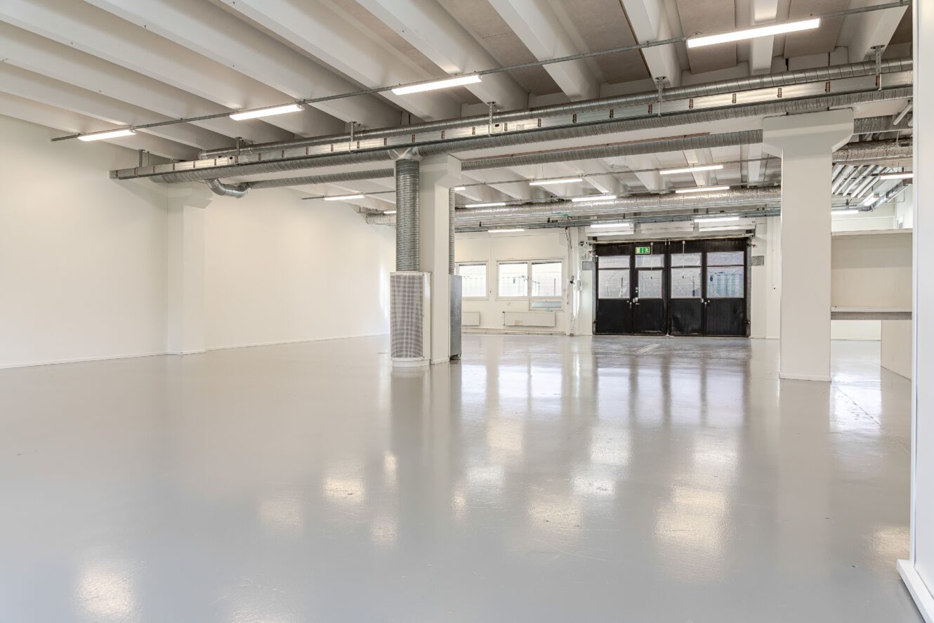 Rymlig, tom industrilokal med polerade golv, vita väggar, synliga rör i taket, stora fönster och svarta dubbeldörrar i bakgrunden.