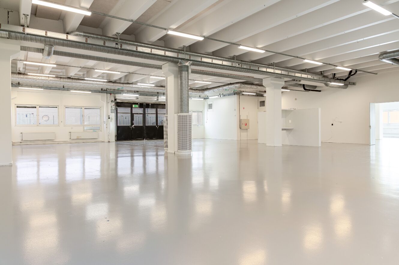 Rymlig, tom industrilokal med vita väggar, stora fönster, synliga rör och polerat betonggolv under fluorescerande taklampor.
