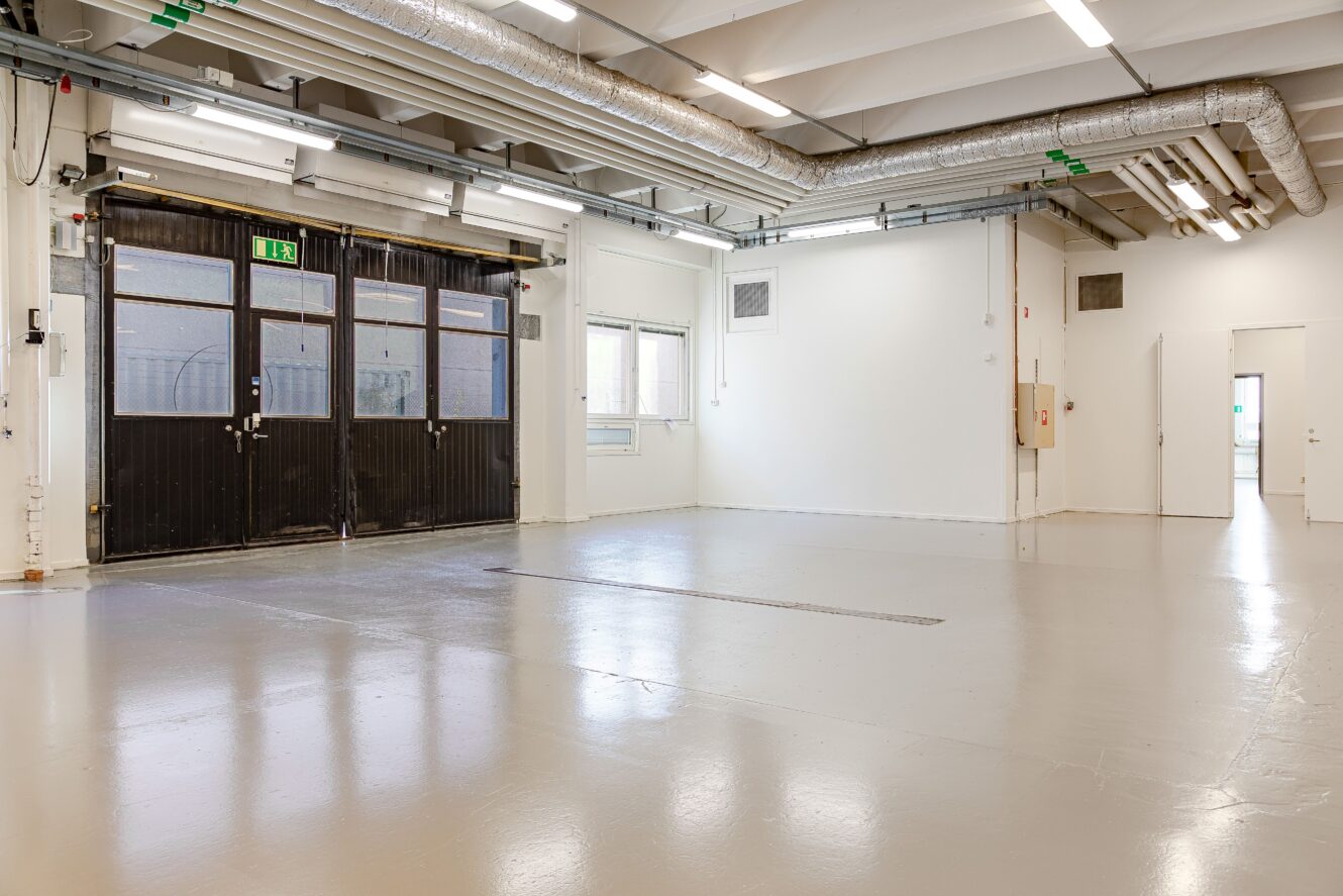Tomt industriellt lagerutrymme med polerade betonggolv, synliga takrör, stora fönster och en dubbel garageport.