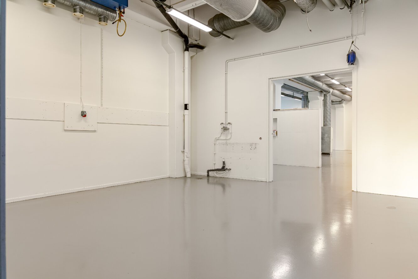 Tomt industrirum med polerat grått golv, vita väggar, synliga rör och ventilationskanaler i taket, anslutet till ett liknande intilliggande utrymme.