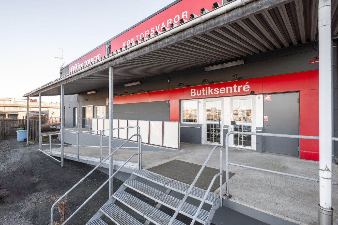 En kommersiell byggnads exteriör med en metalltrappa som leder till glasdörrar märkta Butiksentré under en röd skylt.