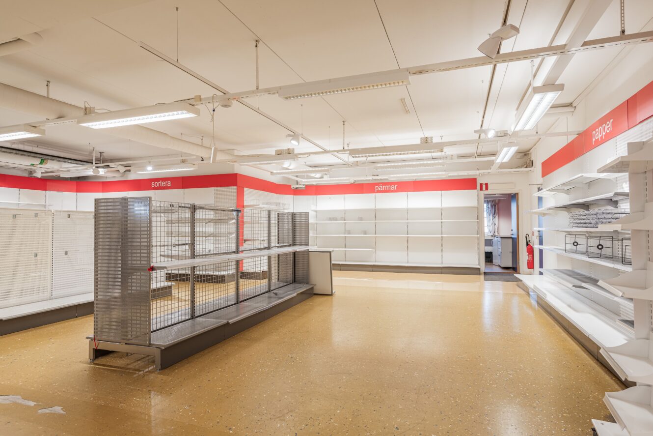 En tom butiksinredning med tomma hyllor och skyltställ, under starka taklampor och röda avdelningsskyltar på väggarna.