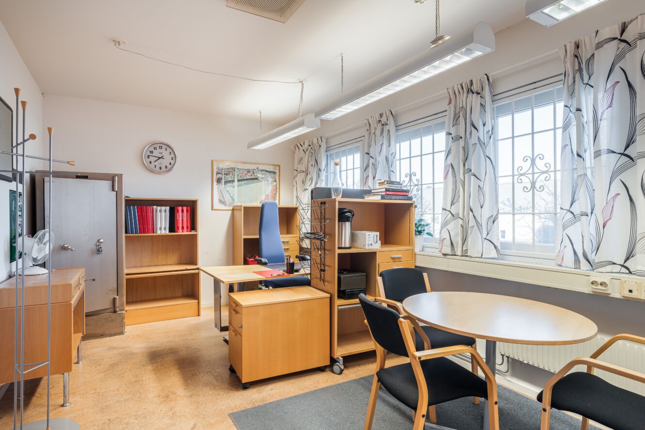 Ett prydligt kontor med trämöbler, två skrivbord med stolar, ett runt mötesbord med stolar, hyllor med pärmar, en klädhängare, en väggklocka och fönster med mönstrade gardiner.