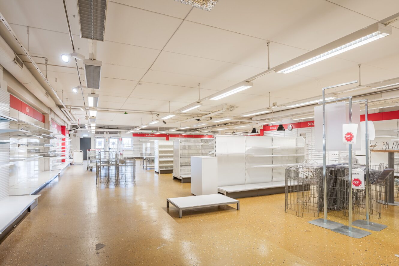 Nästan tom butiksinredning med tomma hyllor, ställningar och minimalt med varor kvar, i starkt fluorescerande belysning.