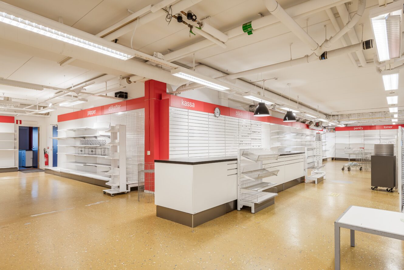 En starkt upplyst, tom butik med vita hyllor, diskar och röda skyltar; utrymmet verkar rent och tomt på varor.