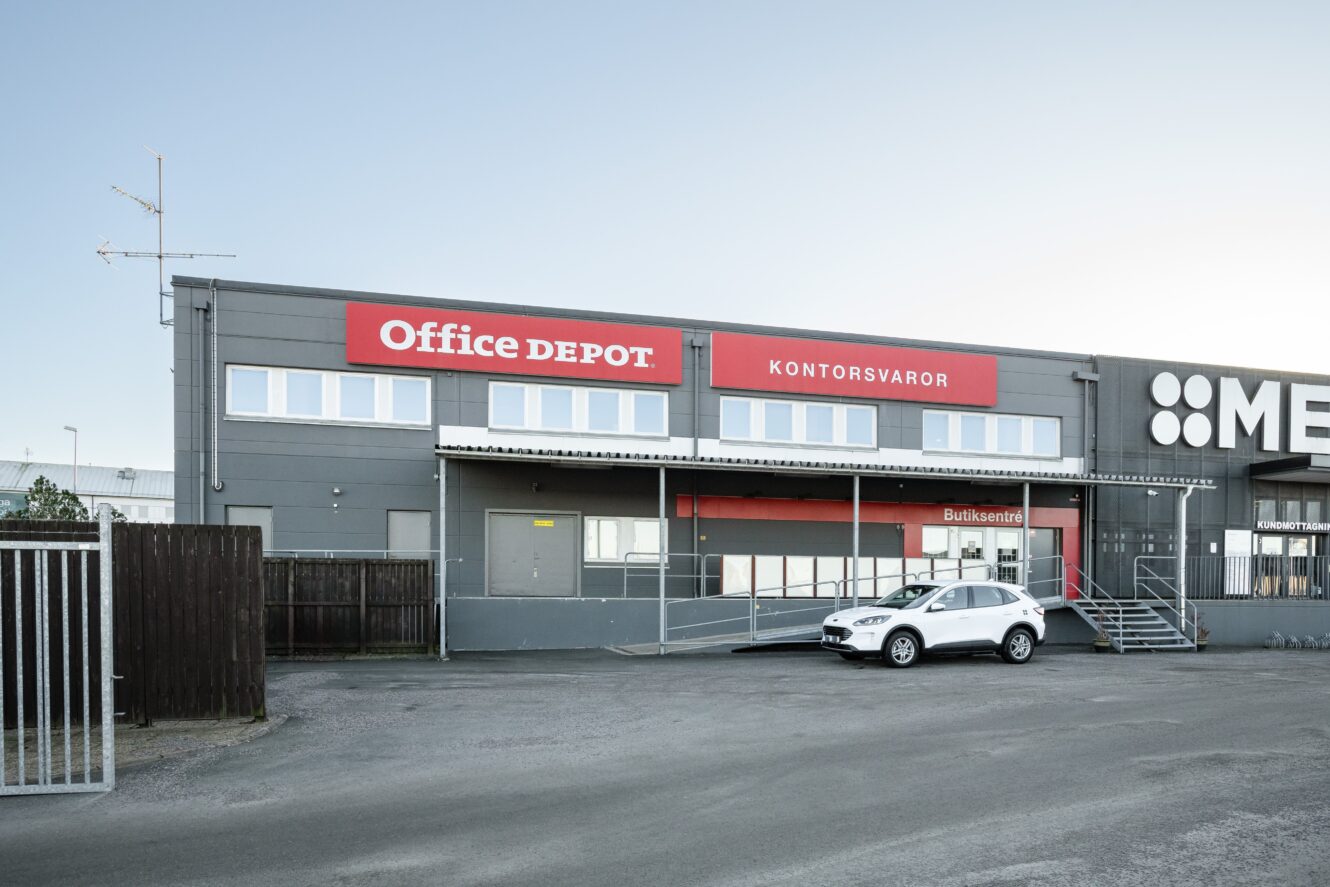 En grå kontorsvaruhusbyggnad med en röd Office Depot-skylt, märkt KONTORSVAROR, och två vita bilar parkerade framför vid en ramp och trappa.