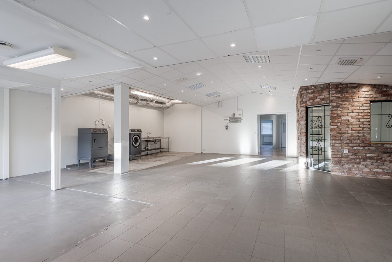 Stort, tomt industrirum med klinkergolv, synliga kanalsystem, två industritvättmaskiner, en accentvägg i tegel och stark takbelysning.