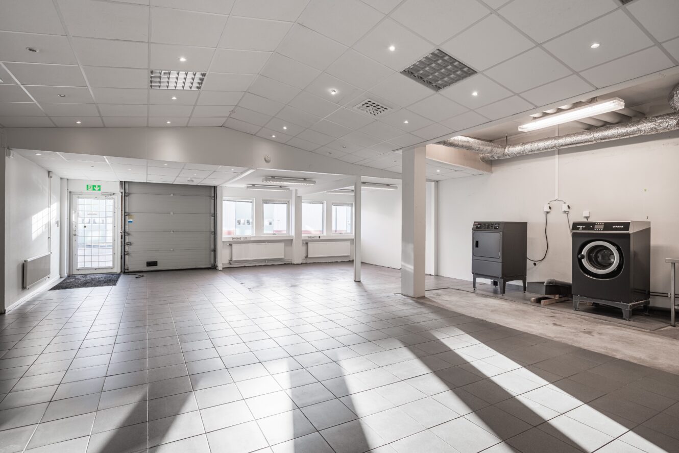 En rymlig, kaklad industrilokal med en garageport, flera fönster och två stora tvättmaskiner längs den högra väggen.