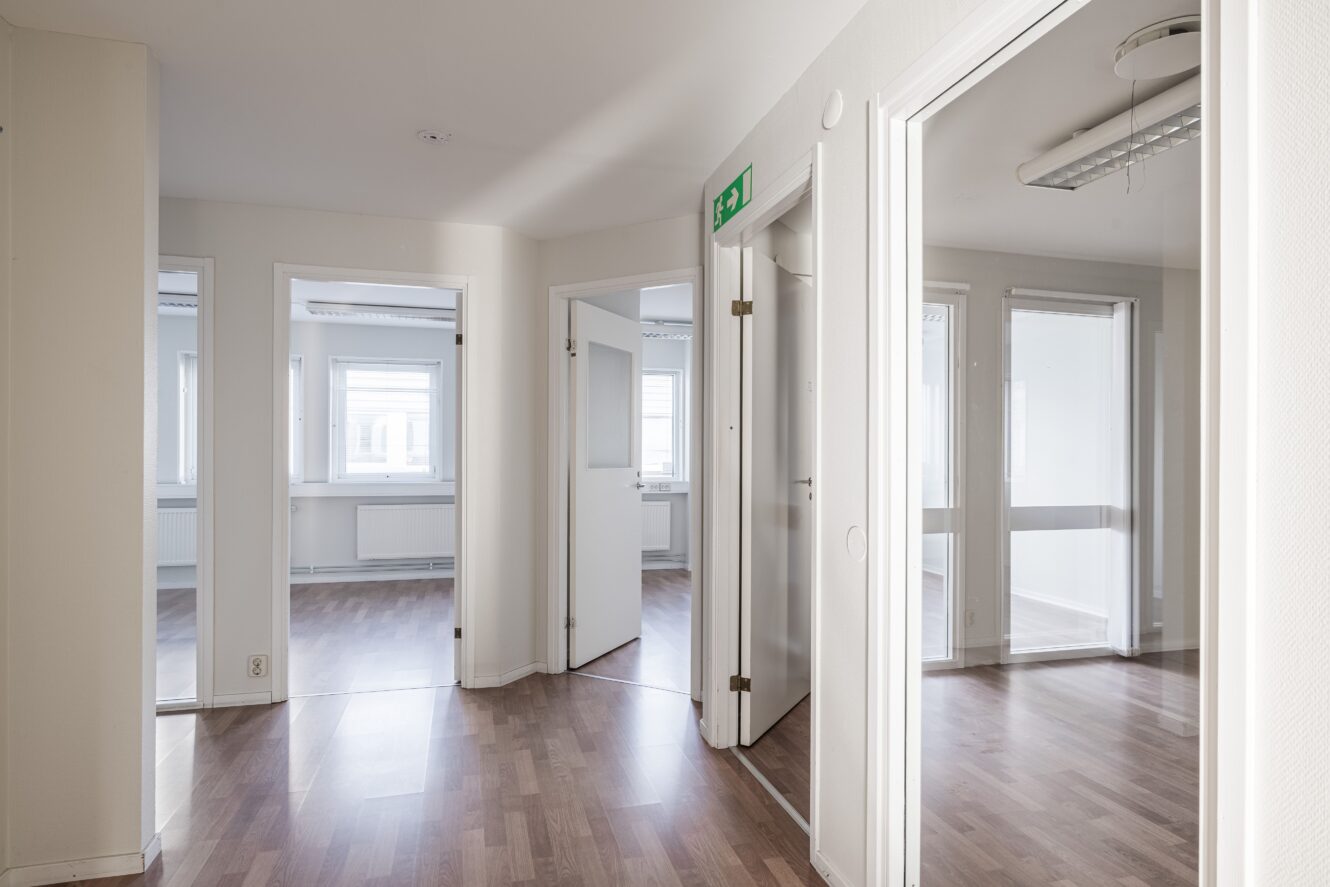 En ren, tom korridor med vita väggar och dörrar som leder till flera omöblerade rum, med trägolv och en nödutgångsskylt på väggen.