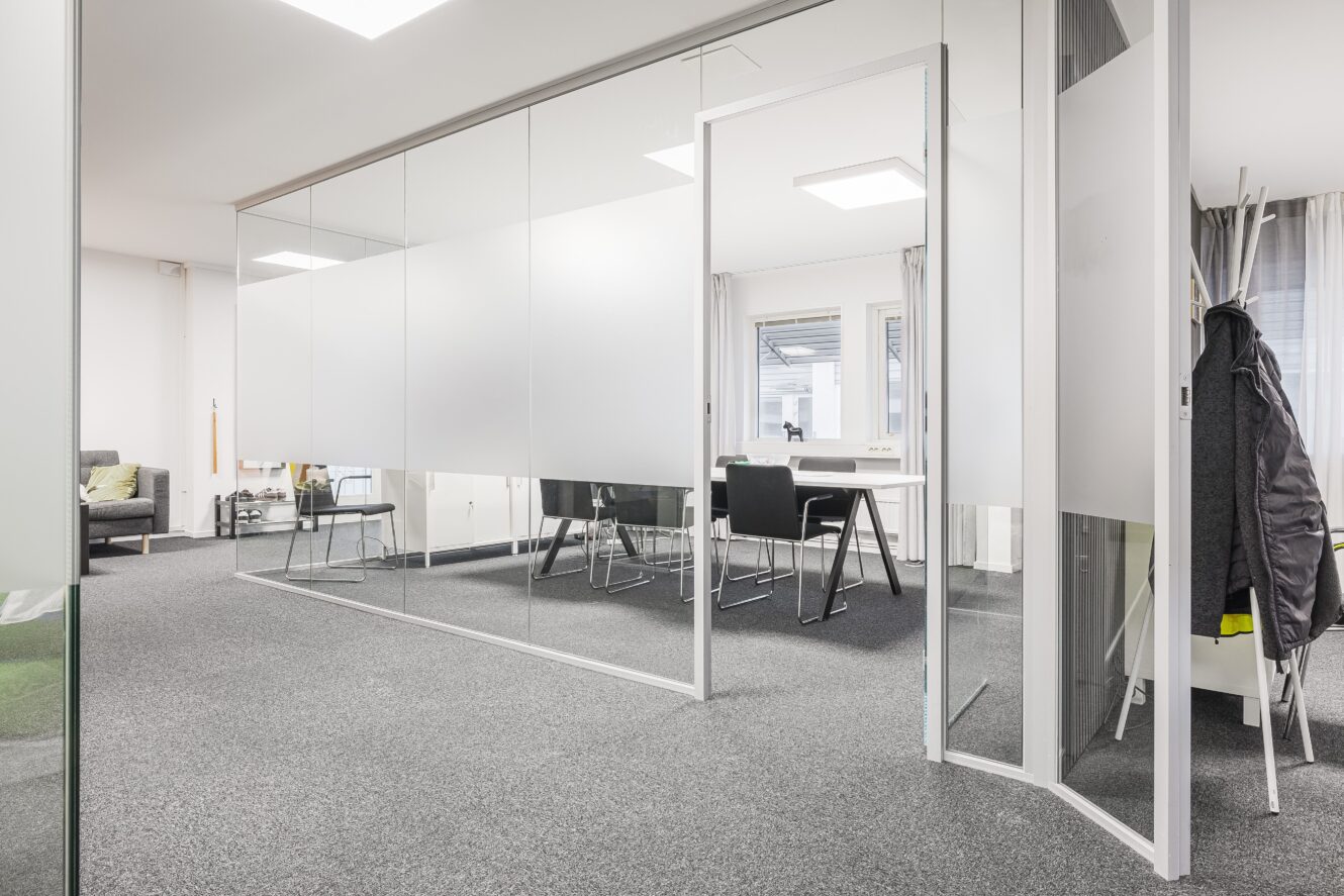 Modernt kontor med mötesrum med glasväggar, svarta stolar runt ett vitt bord och en kappa hängande på en hylla; öppen dörr leder till huvudkontoret.