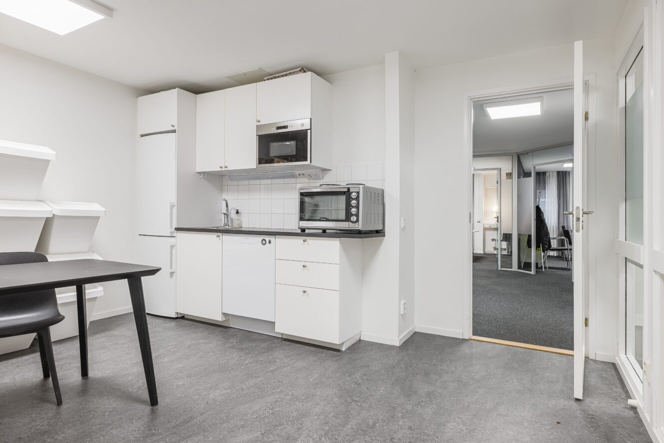 Modernt pentry med vita skåp och vitvaror som kylskåp, mikrovågsugn och brödrost; intilliggande rum med kontorsstolar som syns genom en öppen dörr.