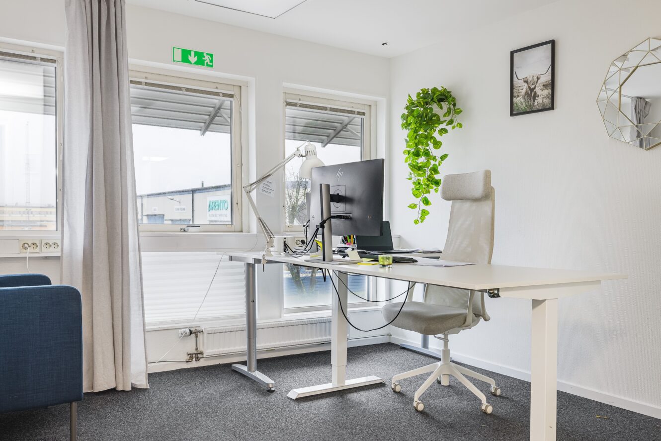 Ett modernt kontor med skrivbord, dator, kontorsstol, lampa, växt, väggkonst och ett fönster som visar en industribyggnad utanför.