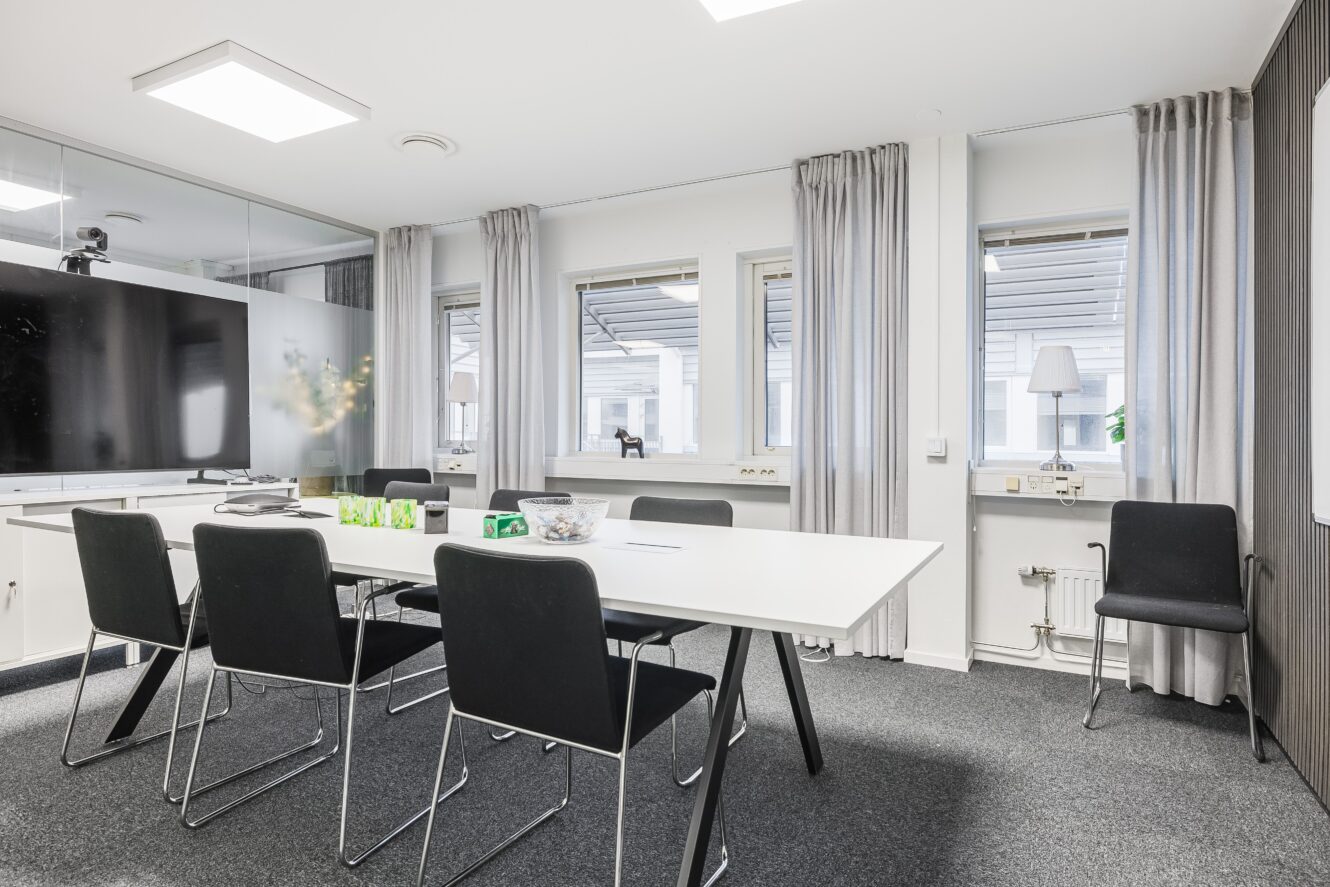 Ett modernt konferensrum med ett vitt bord, sex svarta stolar, en stor väggmonterad skärm och tre fönster med grå gardiner.