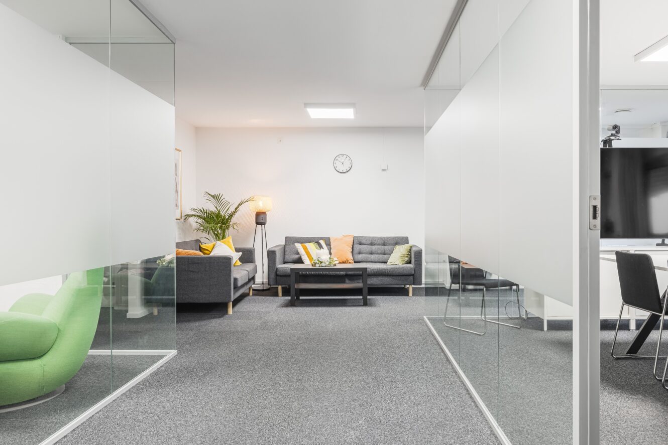 Modern kontorslounge med grå soffor, en grön fåtölj, ett soffbord, en inomhusväxt och en väggklocka. Glaspartier skiljer loungen från ett intilliggande mötesområde.