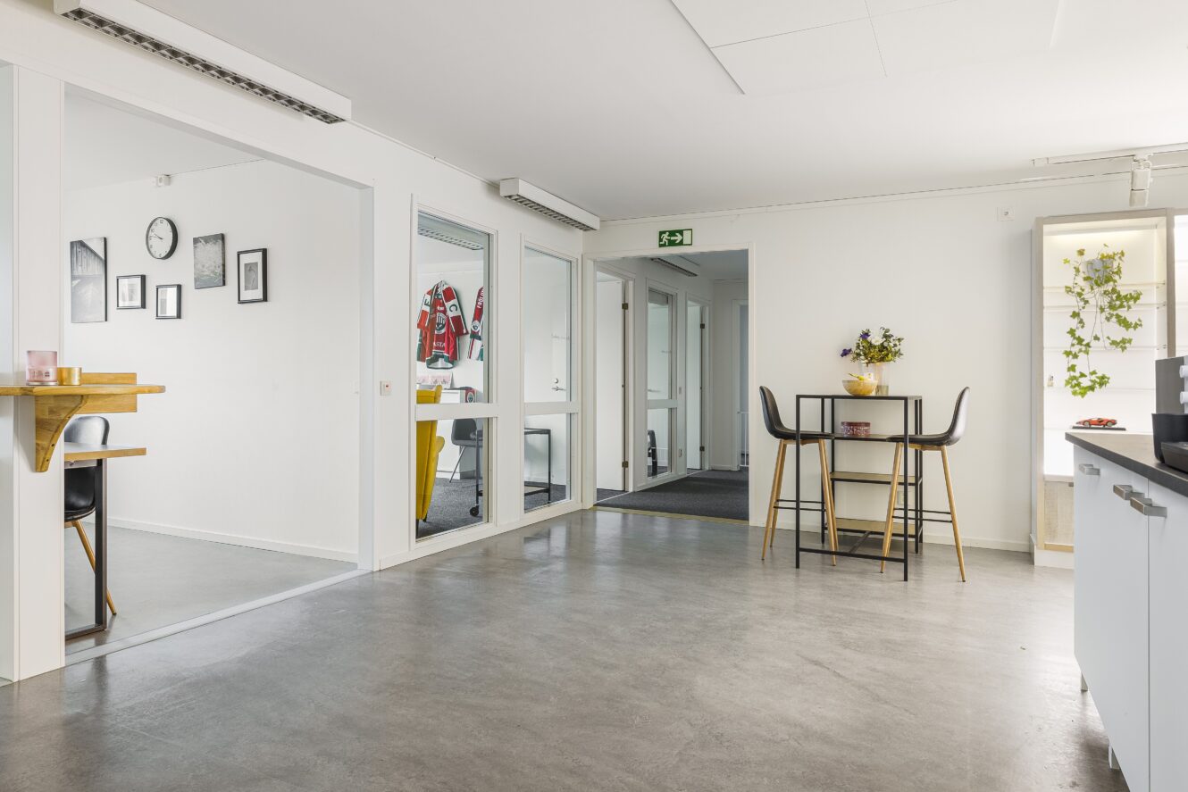 Modernt kontorskök med ett litet matbord och stolar, betonggolv, vita väggar och synlig hall och angränsande rum.