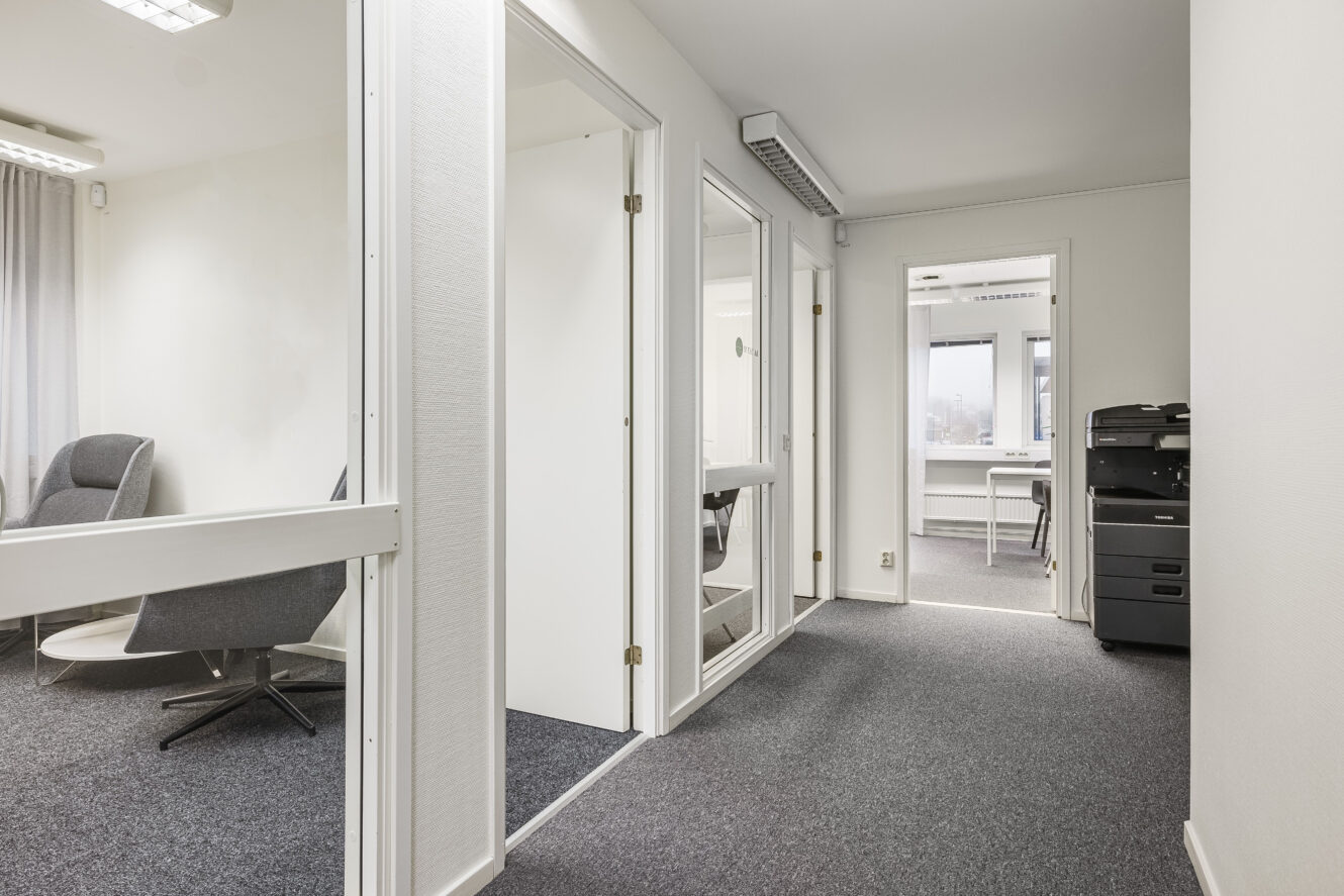 En modern kontorskorridor med vita väggar, grå heltäckningsmatta, glasdörrar, en sittgrupp till vänster och en skrivare vid ett fönster i slutet av korridoren.