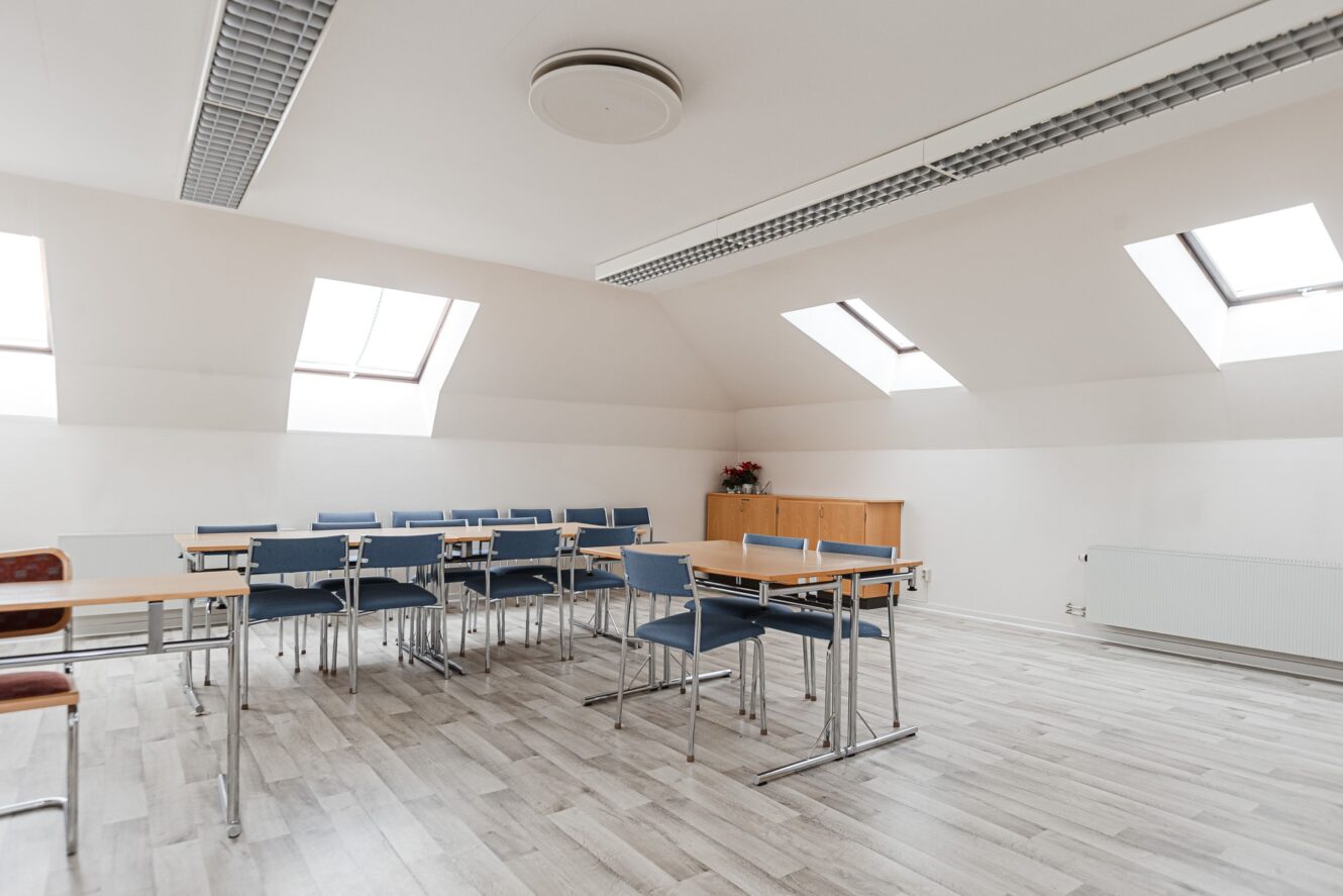 Ett ljust, tomt klassrum med blå stolar, träbänkar, ett ljust trägolv och stora takfönster som släpper in naturligt ljus.