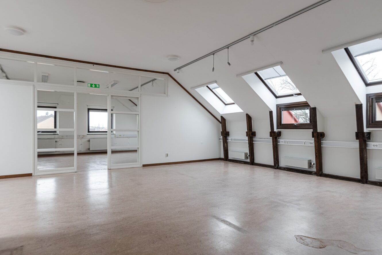 Tomt rum med snedtak, stora fönster med takfönster och en glasvägg; synliga värmeenheter längs väggen och en liten köksdel i bakgrunden.
