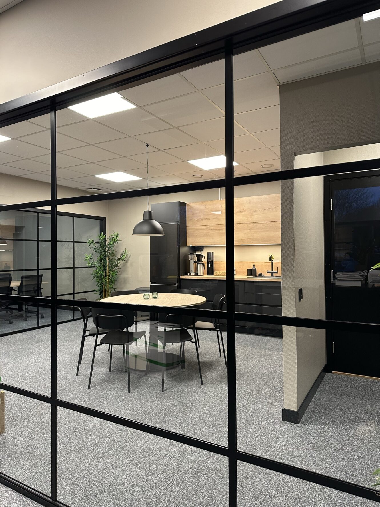 Modernt kontorskök och mötesområde med ett runt bord, fyra stolar, pendellampa och pentry, sett genom glasväggar.