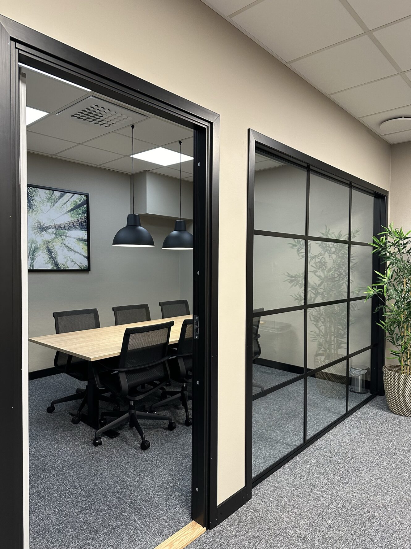 Modernt mötesrum med träbord, svarta kontorsstolar, pendellampor, inramade konstverk och glasvägg; krukväxt i korridoren.