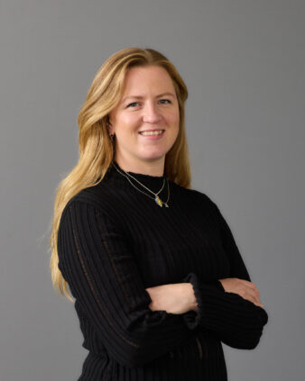 En kvinna med långt blont hår och svart långärmad tröja står med armarna i kors och ler mot en grå bakgrund.