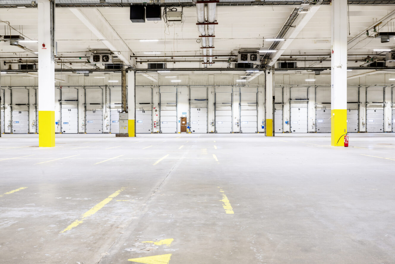 Interiör av en tom lagerlokal med betonggolv, gula markeringar, vita väggar, lastkajsportar och stödpelare med gula baser.