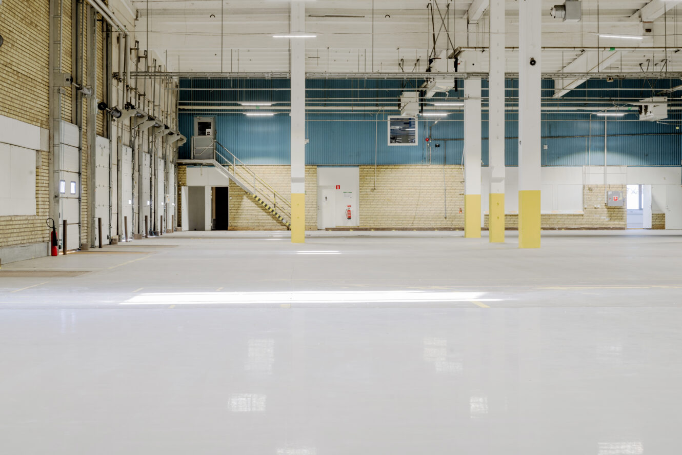 Interiör av ett tomt industrilager med vita golv, pelare, lastkajer och en trappa som leder till en sektion med blå väggar.