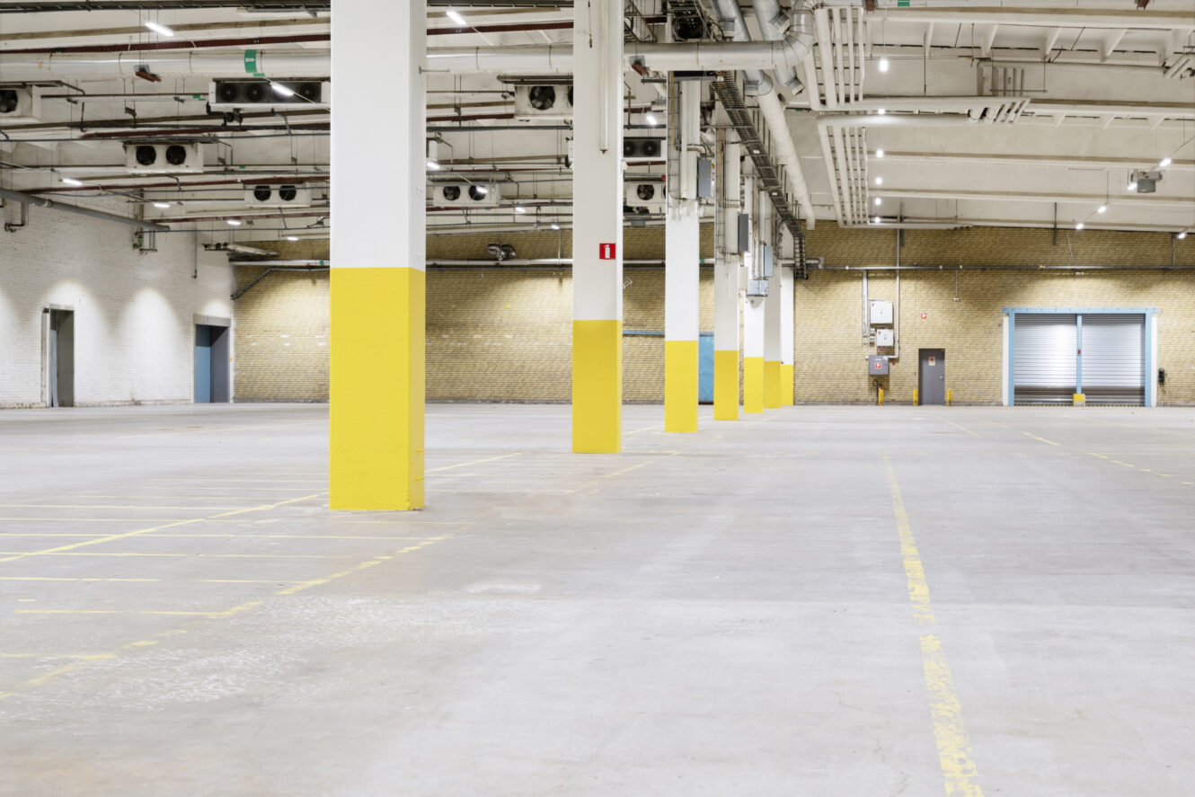 Tom lagerlokal med gulmålade stödpelare, tegelväggar, synliga rör i taket och markerade parkeringsplatser på betonggolvet.