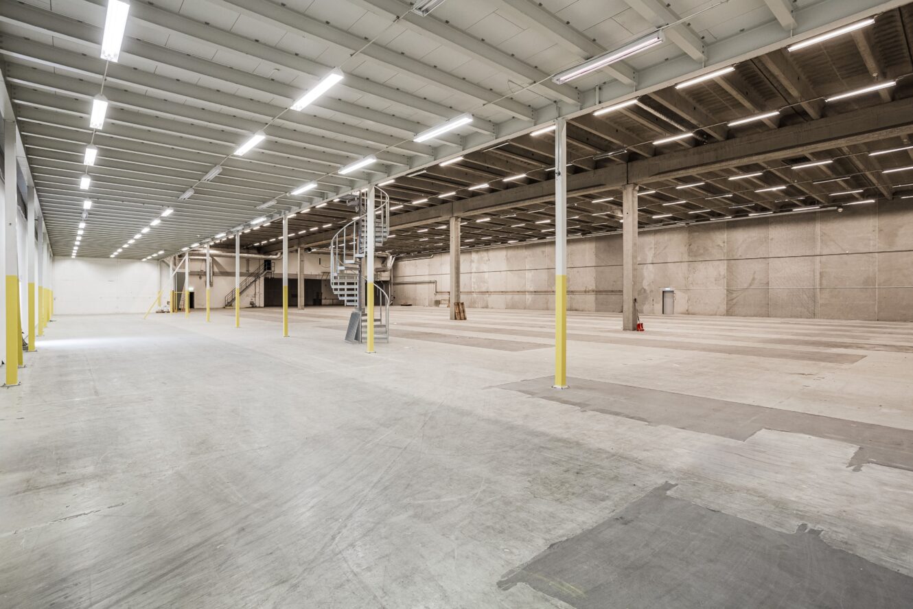 Stor, tom lagerlokal med betonggolv, högt i tak, synliga bjälkar, lysrörsbelysning och en spiraltrappa i metall nära mitten.